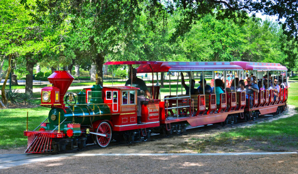 A red and green train with passengers moving through green trees 