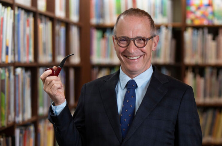 A promotional image of David Sedaris holding a pipe in front of bookshelves