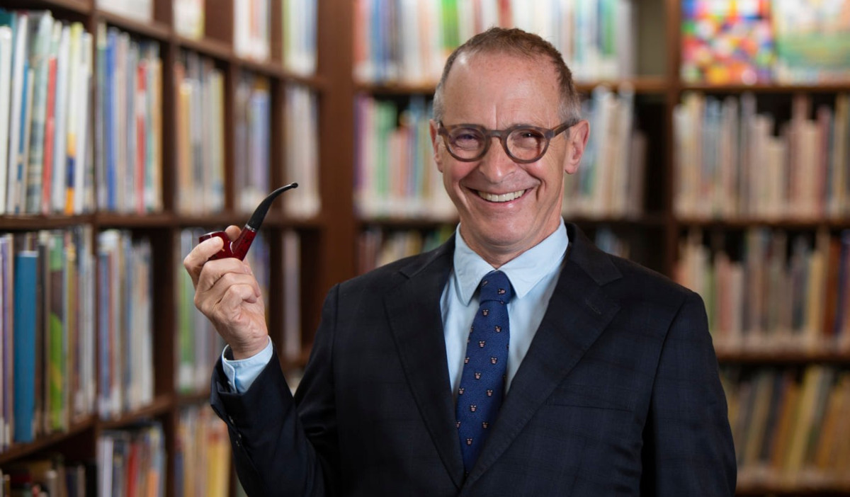 A promotional image of David Sedaris holding a pipe in front of bookshelves