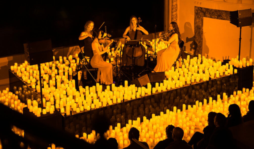 A string quartet performs on stage, surrounded by hundreds of candles