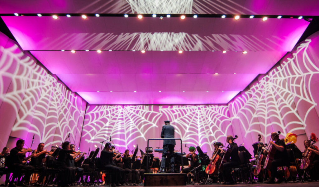 An orchestra performs in front of a projection of white spider webs on a purple wall