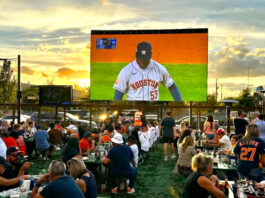 Fans sit at tables in an outdoor patio as an Astros game plays on a big screen