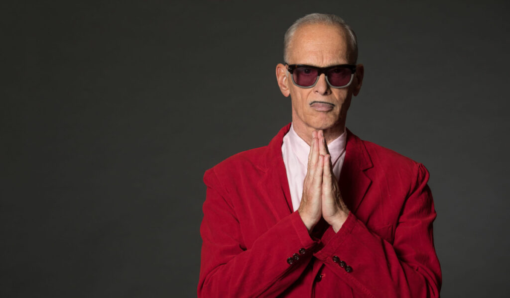 A promotional image of John Waters in a red coat clasping his hands in front of his face