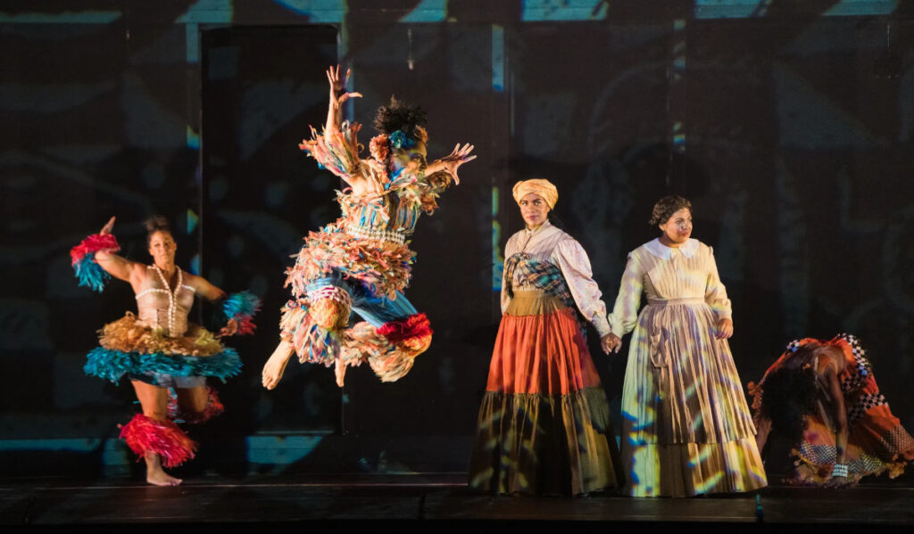 Women in African clothing dance around two actors wearing long dresses