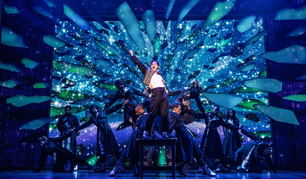 An actor dressed as Michael Jackson performs a dance move in front of blue and green shards behind him