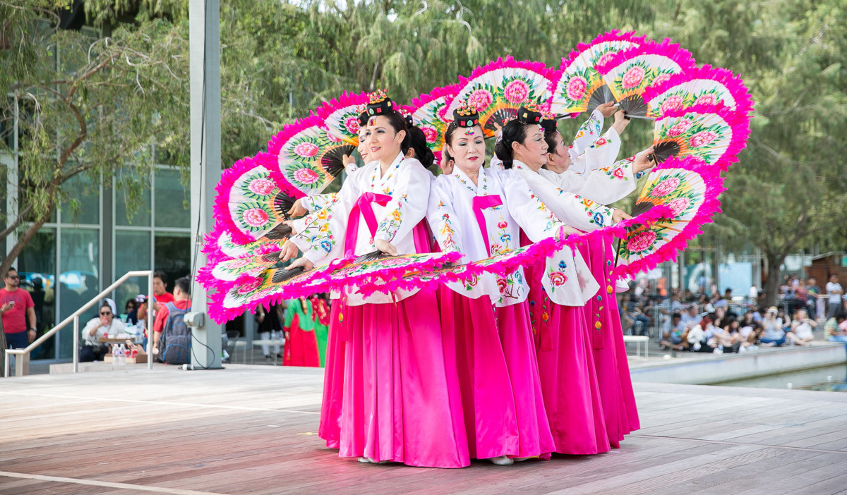 Dancers in pink and white costumes form a circle with pink fans