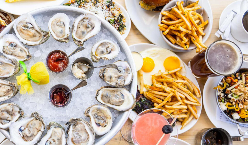An overhead view of a plate of oysters on ice with plates of burgers, eggs, and cocktails