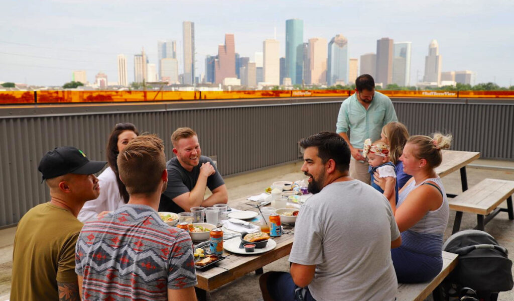 A group of people sitting at a table with food and drink and the Downtown skyline behind them
