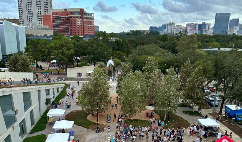 An aerial view of the MFAH campus with crowds of people, white canopy tents, and the skyline of the Medical Center