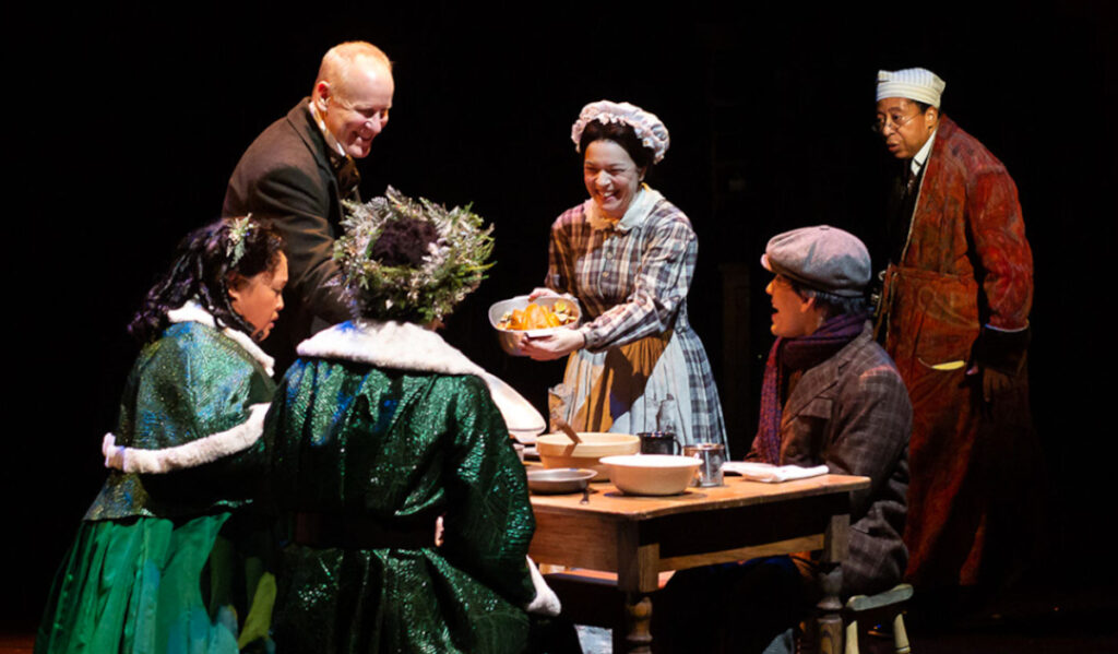 Five people around a dining table as the character Ebenezer Scrooge watches from the background