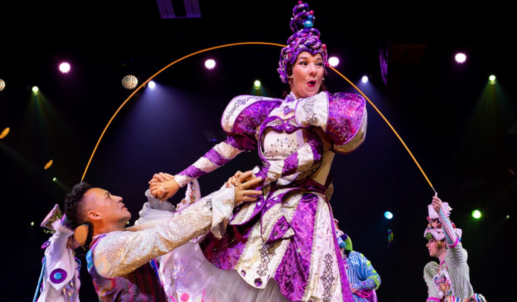 A cirque performer in purple and white costume is hoisted up by another performer