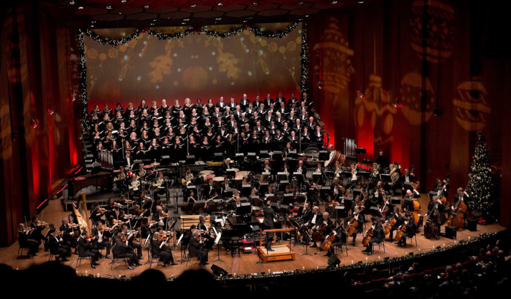 A full orchestra and choir performing on a holiday-decorated stage