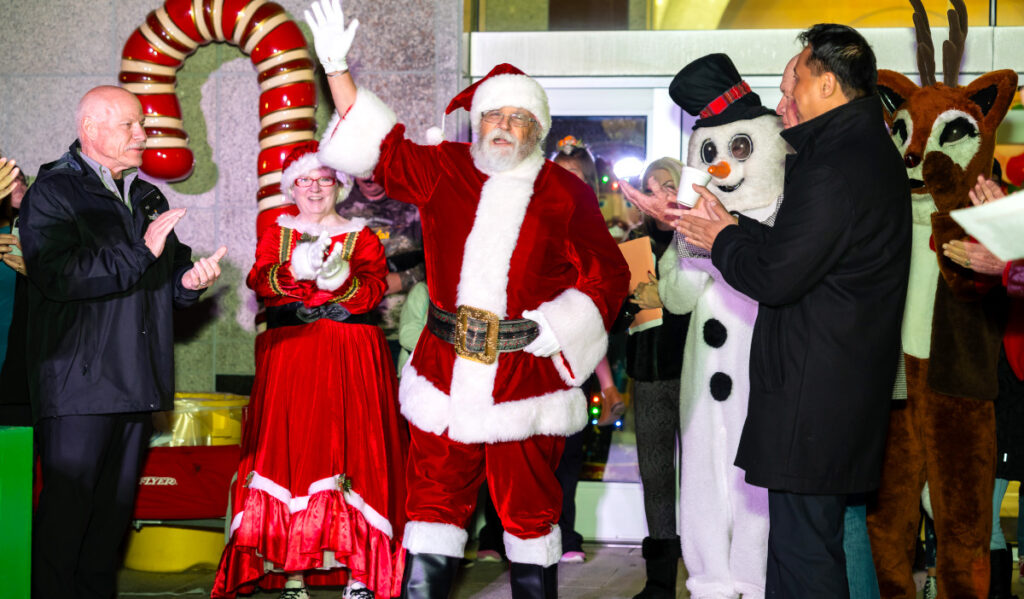 Santa Claus waves at a crowd with Mrs. Claus and a Snowman behind him