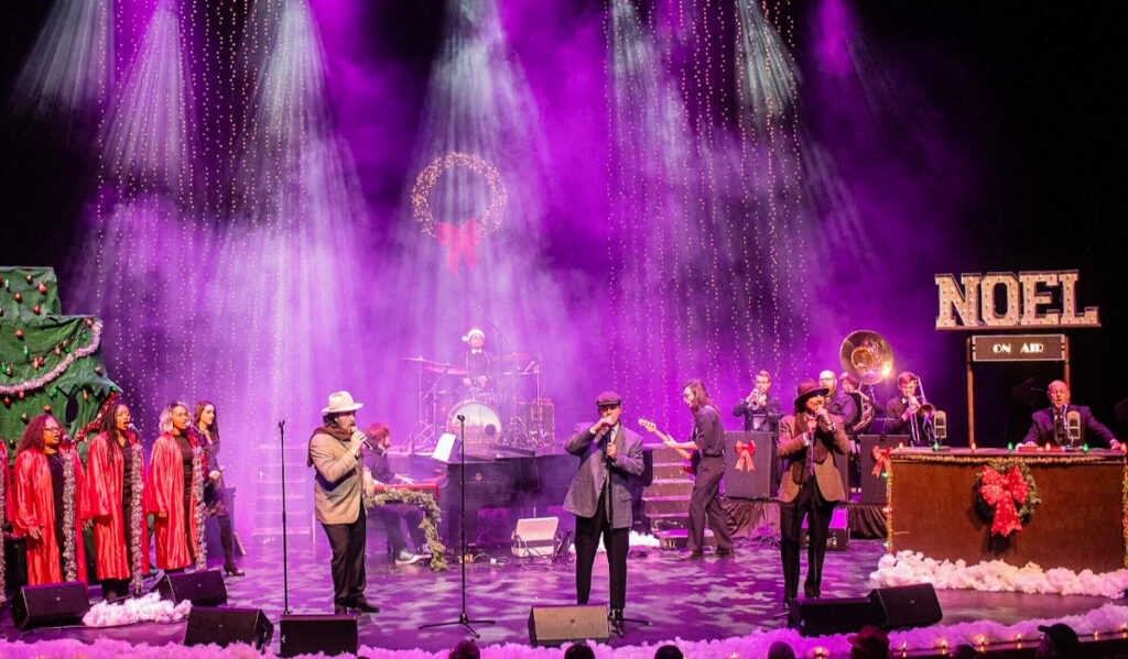 A band performs on a holiday-decorated stage with purple light above them