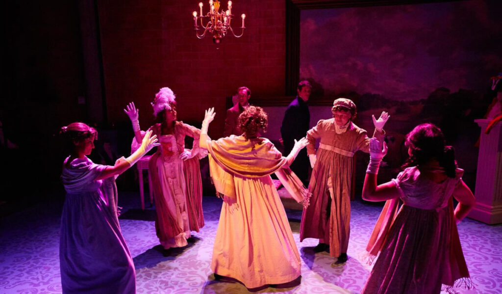 Four people in Victorian dress dance around one person lit in orange light