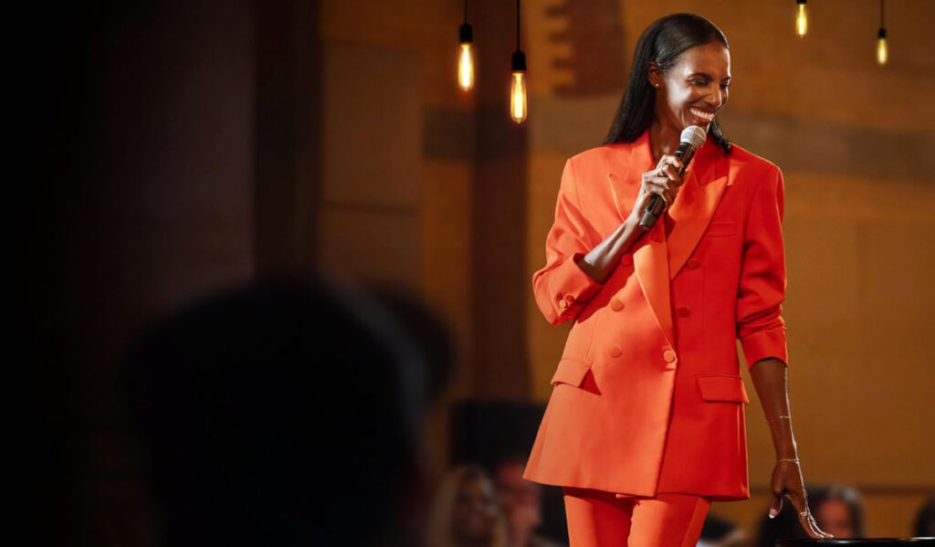 Comedian Zainab Johnson performing on stage in a red suit