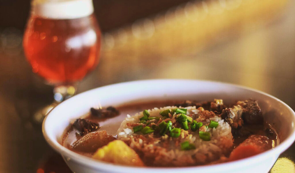 A bowl of beef curry with a glass of beer in the background