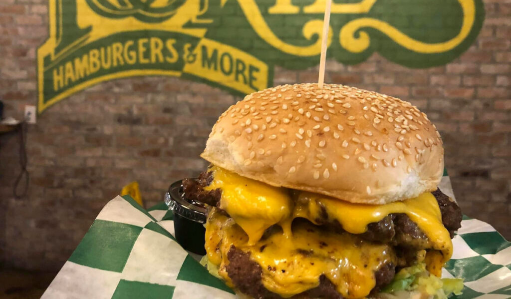 A burger with multiple patties and gooey cheese with a Tookie's sign behind it