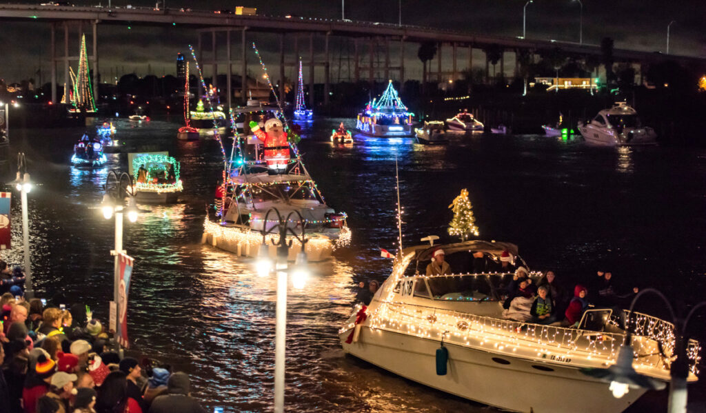 Boats decorated with Christmas lights lined up as a crowd of people watch