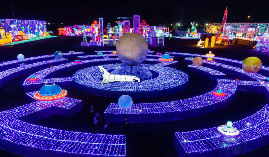 An overhead view of a space themed labyrinth in purple lights with other holiday light attractions in the distance