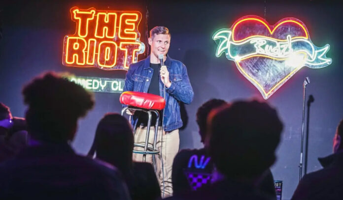 Comedian Brian Gendron performs while holding a stool on stage