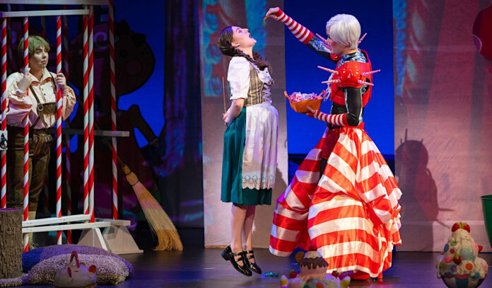 A witch holding a treat above a jumping child's mouth. The child holds a wand behind her back and another child watches from behind bars