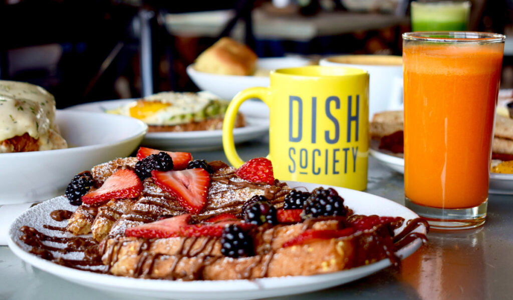 A plate of nutella-drizzled french toast with a glass of orange drink and a mug that says Dish Society