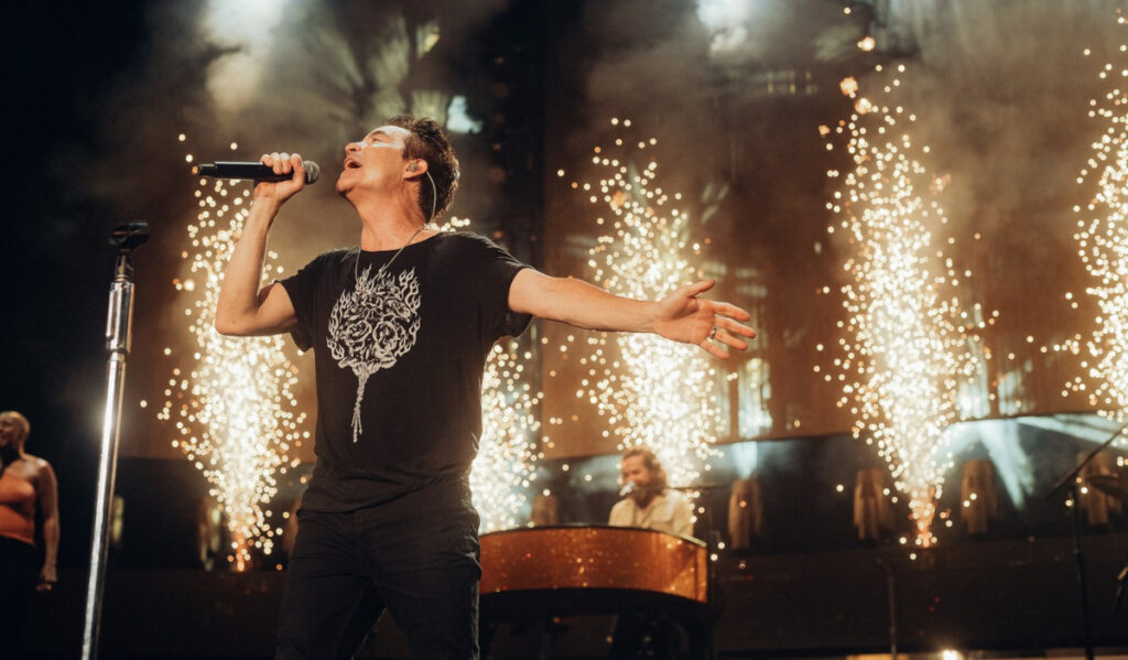 A singer performs while outstretching one hand, with sparks behind him