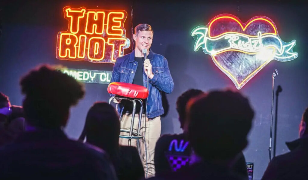 Comedian Brian Gendron performs while holding a stool on stage