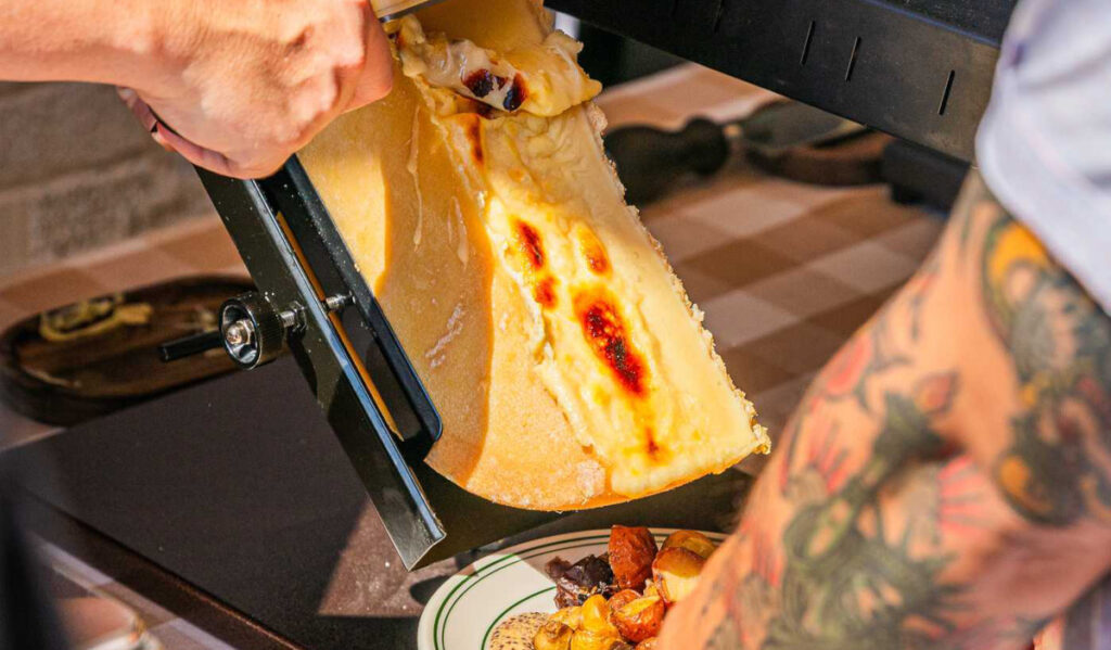 A hand scraping melted raclette cheese from a cheese wheel onto a plate of breads