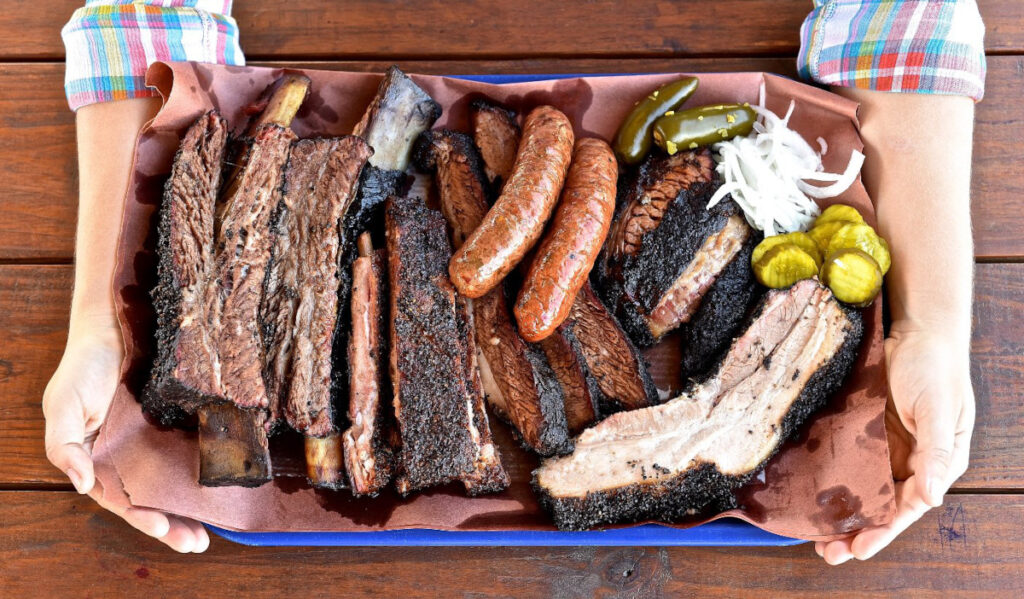 A tray full of barbecue ribs, sausage and other meats, held by two hands