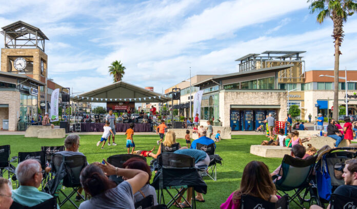 Kids playing on a grass field at a shopping center while adults sit in lawn chairs nearby