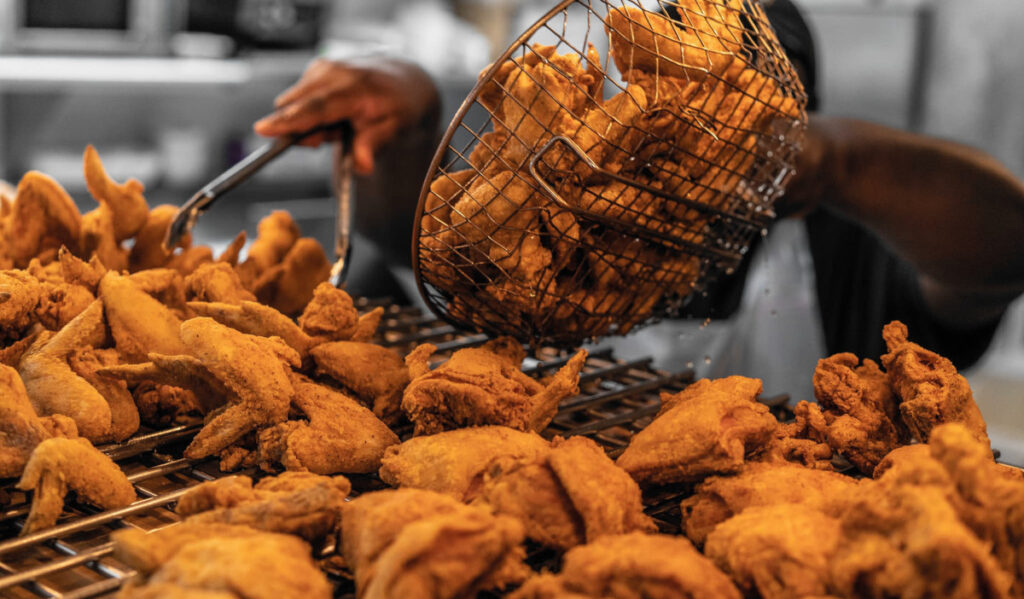 A person dumps a basket of fried chicken onto a grate filled with fried chicken pieces