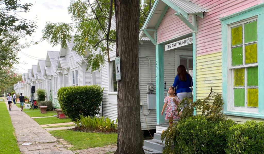 A child and adult standing in the doorway of a house that is part of a row of shotgun-style homes painted white