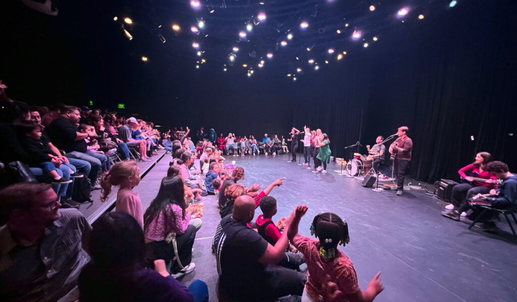 Families sit in a theater as a group of musicians perform and interact with the crowd