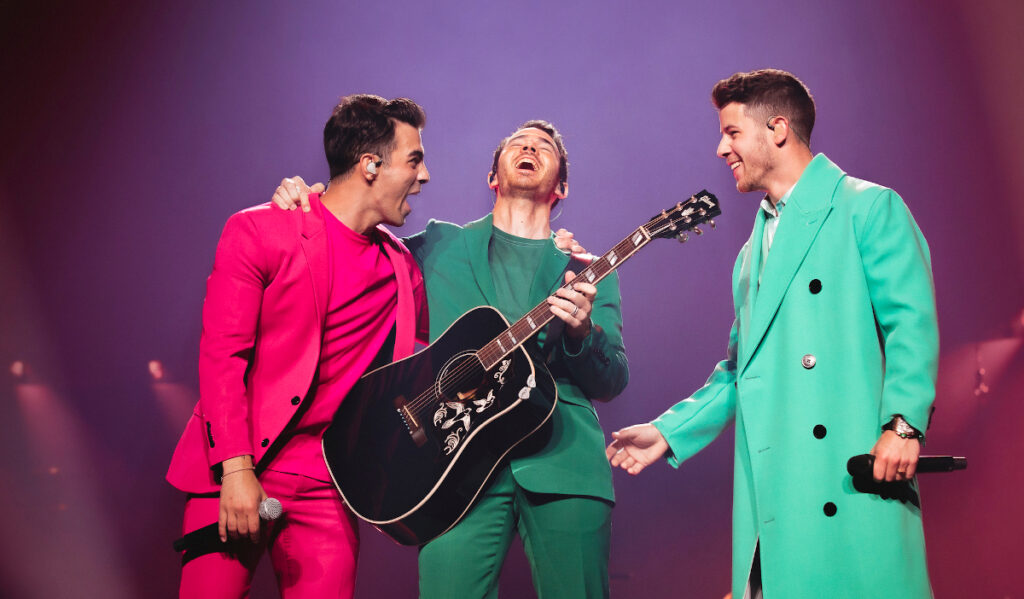 The Jonas Brothers, wearing pink, green and teal suits respectively, laugh on stage 