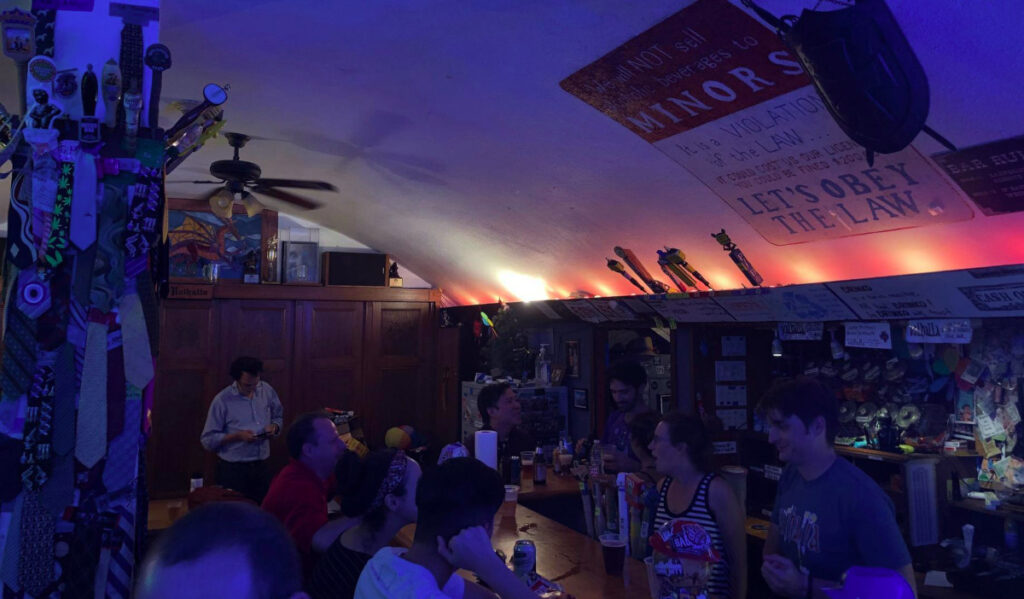 A dark, blue-lit interior of a small bar with neckties taped to a central column