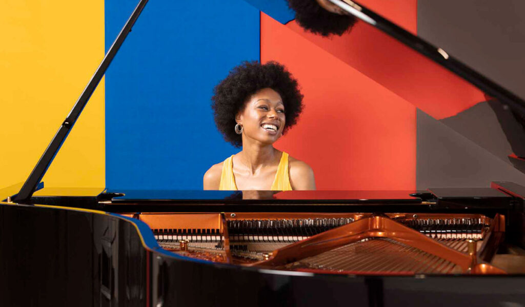 Isata Kanneh-Mason sits behind a piano smiling with yellow, blue, red and brown wall behind her
