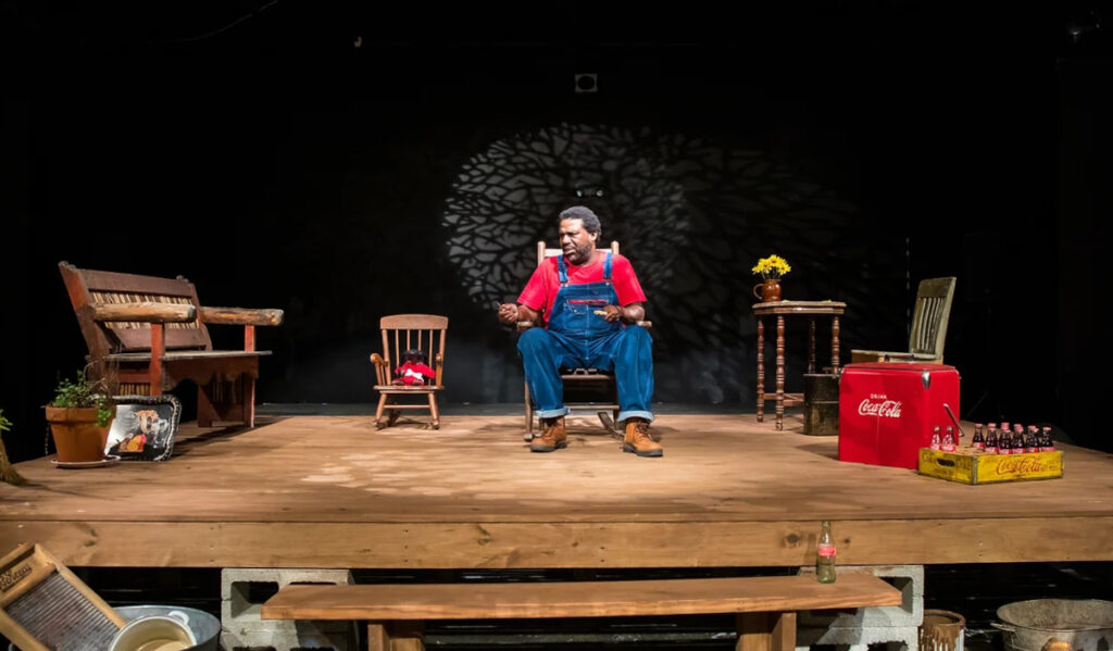 A man in overalls and a red shirt sits in the middle of a stage surrounded by living room furniture
