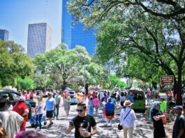 A bustling, sunny scene with people walking along streets past art cars and Downtown Houston in the background