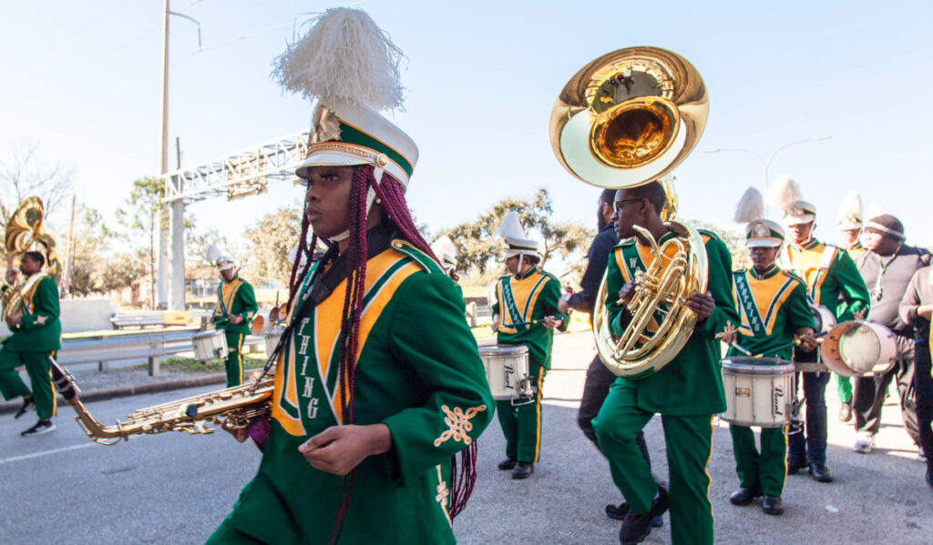 Marching band members walking down the street while holding a saxophone, tuba and drums