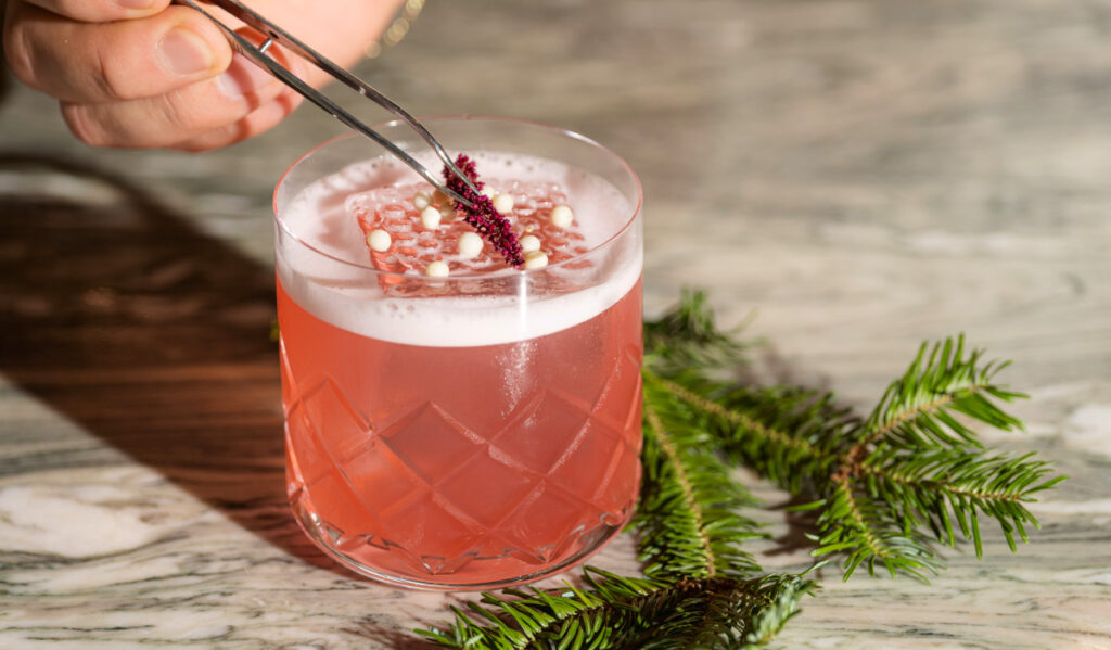 A hand with tweezers places a garnish on top of a pink cocktail