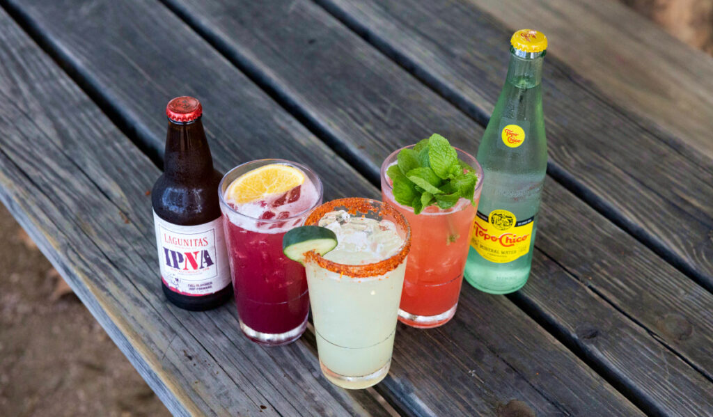 Three cocktails with garnishes on a bench next to a Topo Chico and a Lagunitas IPNA