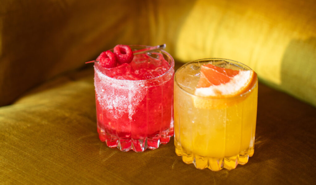 A short red cocktail with berry garnish next to a short yellow cocktail with orange slice