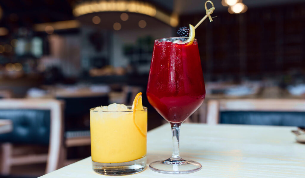 A short, yellow cocktail next to a red cocktail in a stem glass