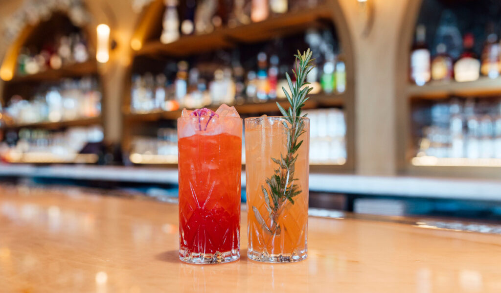 A tall red cocktail next to a tall orange cocktail with a sprig of thyme