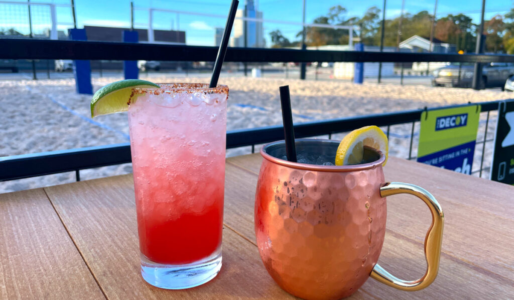 A tall red and white cocktail next to a copper cup with a sand volleyball court in the background