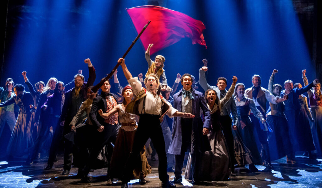 The ensemble cast of "Les Miserables" raises their first with a red flag flying behind them