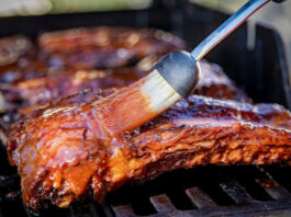 Beef ribs being basted with bbq sauce on a grill with a basting brush