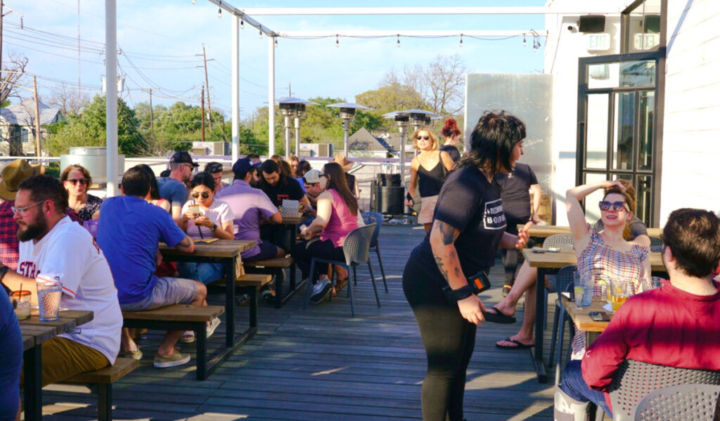 A rooftop patio on a sunny day with people sitting at tables and a server speaking with a customer